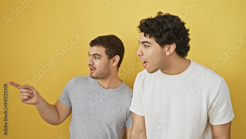 Two hispanic men in casual clothing interact against a vibrant yellow background, the first gesturing while the second watches with surprise. photo
