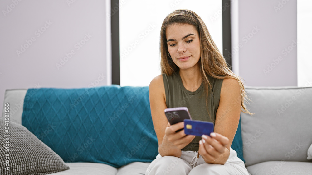Stunning young hispanic woman paying bills with credit card, deeply concentrated on her smartphone, sitting on her cozy sofa, enjoying online shopping in the comfort of her living room Stunning young hispanic woman paying bills with credit card, deeply concentrated on her smartphone, sitting on her cozy sofa, enjoying online shopping in the comfort of her living room