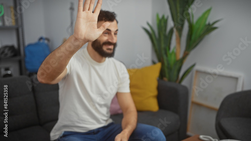 A cheerful man waving in a cozy living room with a white t-shirt, couch, and plant.