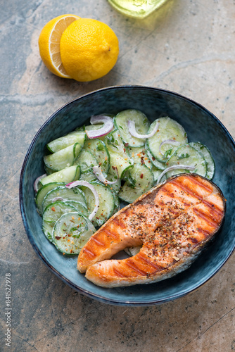 Dark-blue bowl with cucumber salad and grilled salmon steak, vertical shot on a grey and beige granite surface, elevated view