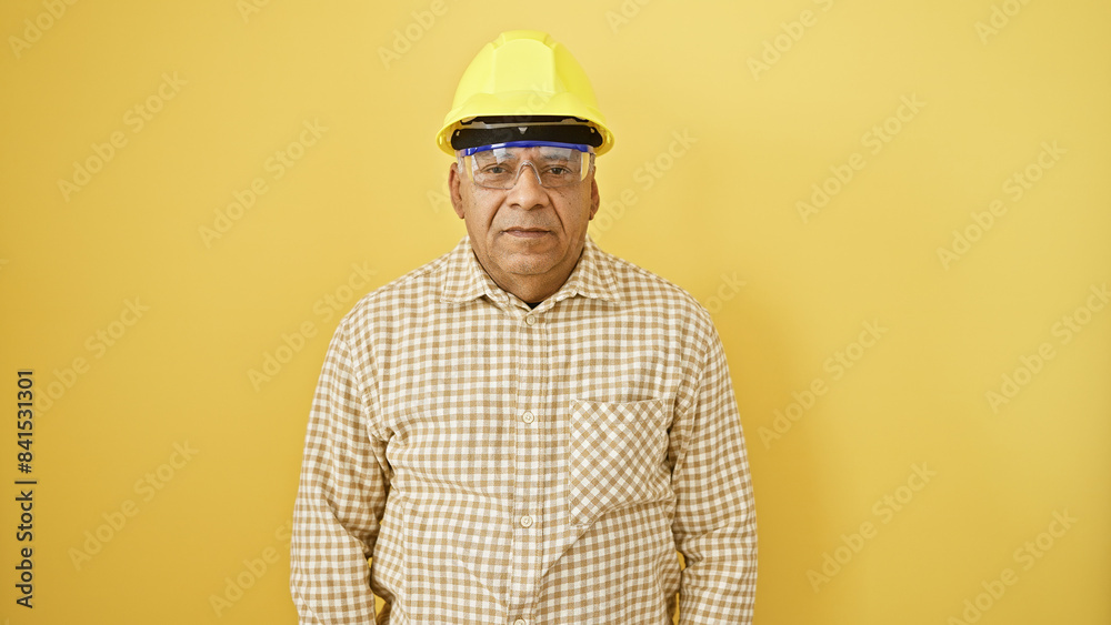 Mature hispanic man wearing a yellow hardhat and safety goggles stands against an isolated yellow background. Mature hispanic man wearing a yellow hardhat and safety goggles stands against an isolated yellow background.