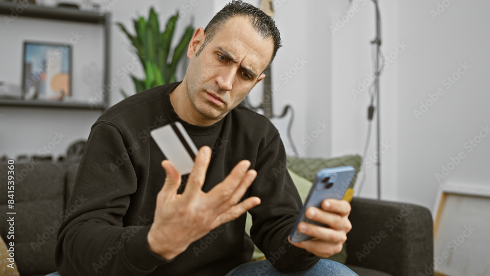 A confused hispanic man examines his credit card while holding a smartphone in a modern living room.
