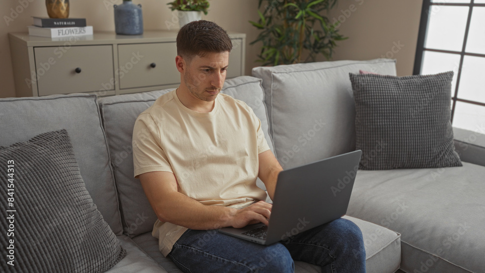 Young hispanic man working on his laptop while sitting on a cozy couch in the living room of his apartment.