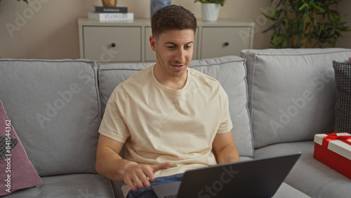 Handsome young hispanic man using a laptop while sitting on a sofa in a cozy living room at home, focusing on the screen with a gift box in the background.