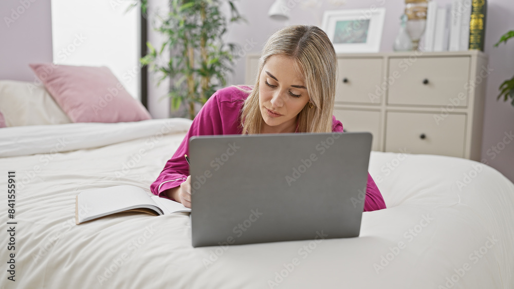 A focused young woman lies comfortably on her bed at home, working on a laptop and taking notes. A focused young woman lies comfortably on her bed at home, working on a laptop and taking notes.