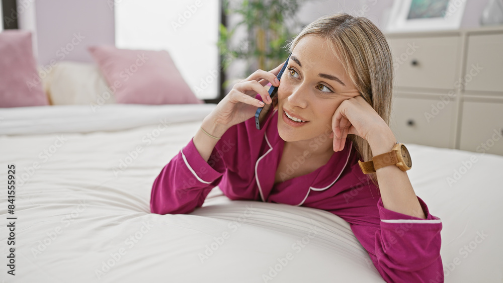 Caucasian woman in pink pajamas talking on a mobile phone while lying on a white bed in a bright bedroom Caucasian woman in pink pajamas talking on a mobile phone while lying on a white bed in a bright bedroom