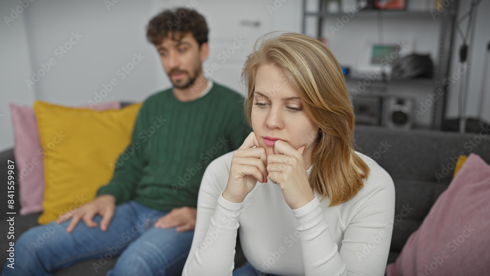 A concerned woman and a contemplative man sit apart on a couch in a modern living room, symbolizing relationship tension.