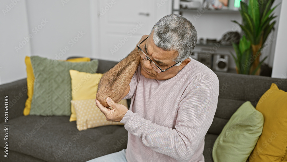 Senior man with glasses holding elbow in pain while sitting on a sofa in a home interior. Senior man with glasses holding elbow in pain while sitting on a sofa in a home interior.