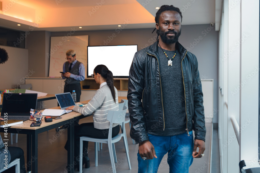 A African Man in a black jacket next to the window in the office and behind him are people working