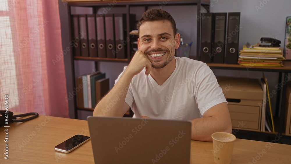 A handsome young hispanic man with a beard smiles confidently in an office setting with shelves full of binders and files in the background. A handsome young hispanic man with a beard smiles confidently in an office setting with shelves full of binders and files in the background.