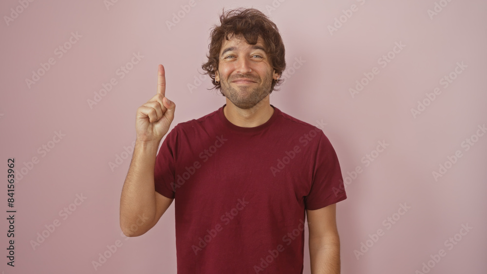 A cheerful young hispanic man with a beard pointing up against a pink isolated background, wearing a casual burgundy shirt. A cheerful young hispanic man with a beard pointing up against a pink isolated background, wearing a casual burgundy shirt.
