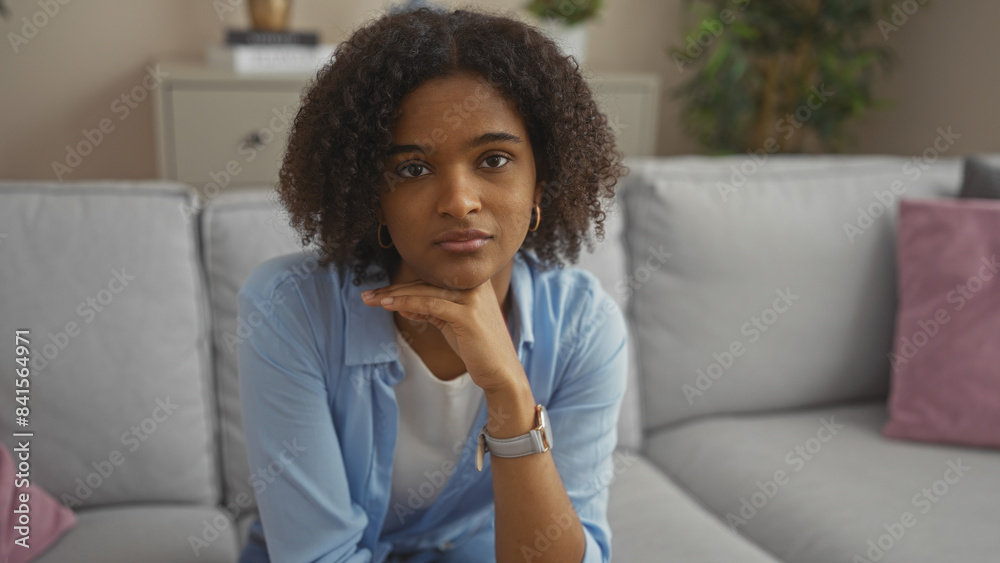 Young african american woman with curly hair sitting on a sofa in a cozy living room, looking thoughtful indoors. Young african american woman with curly hair sitting on a sofa in a cozy living room, looking thoughtful indoors.