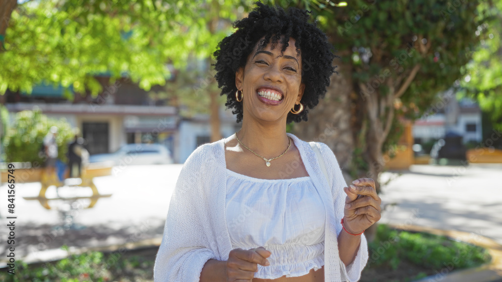 Fototapeta premium A beautiful young african-american woman with curly hair smiles brightly while standing outdoors in an urban park.
