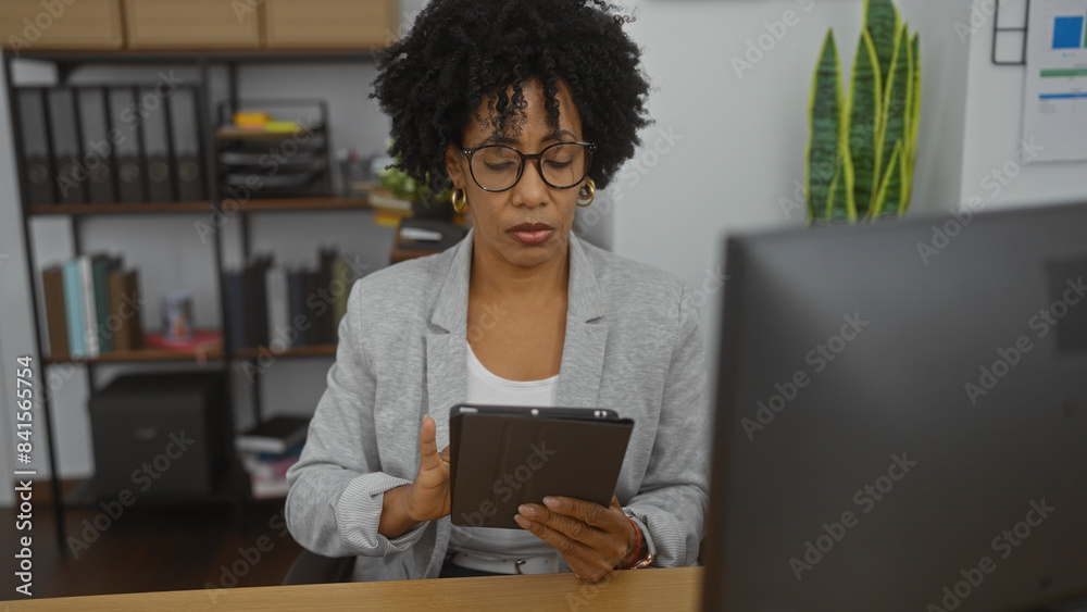 Young african american woman with curly hair in a grey blazer working indoors at an office desk using a tablet with a focused expression and shelves in the background. Young african american woman with curly hair in a grey blazer working indoors at an office desk using a tablet with a focused expression and shelves in the background.