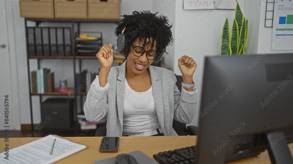 Happy african american woman with curly hair wearing headphones, enjoying music in a modern office workspace with shelves and documents behind her, expressing excitement in front of a computer.