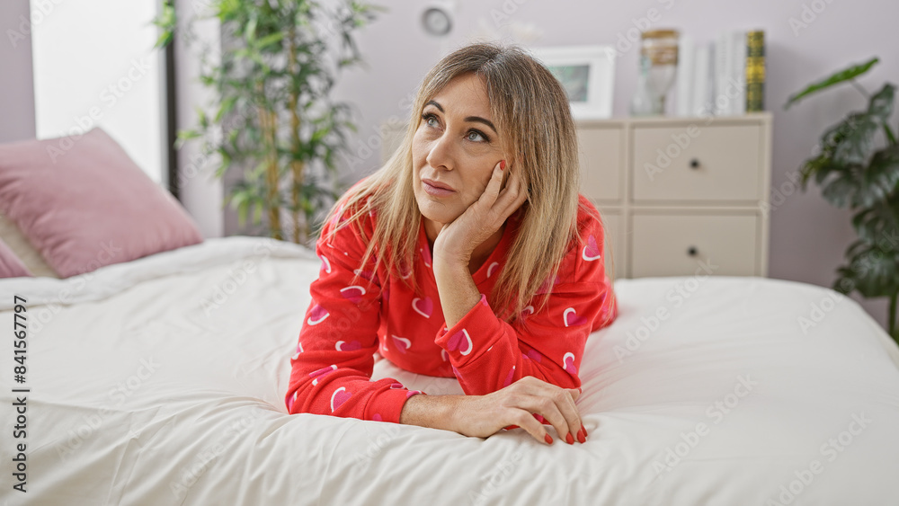 Thoughtful blonde woman in red pajamas lying on a bed inside a cozy bedroom, looking away pensively.