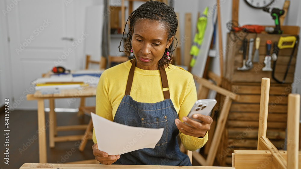 African american woman reviews plans in carpentry workshop with phone in hand African american woman reviews plans in carpentry workshop with phone in hand