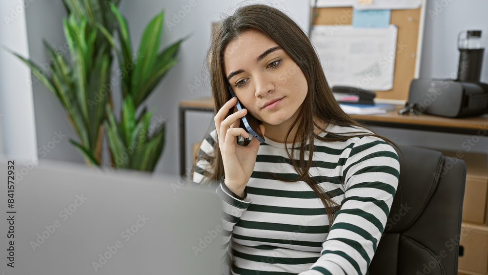 A young latina woman in a striped shirt talking on a smartphone in a modern office environment. A young latina woman in a striped shirt talking on a smartphone in a modern office environment.
