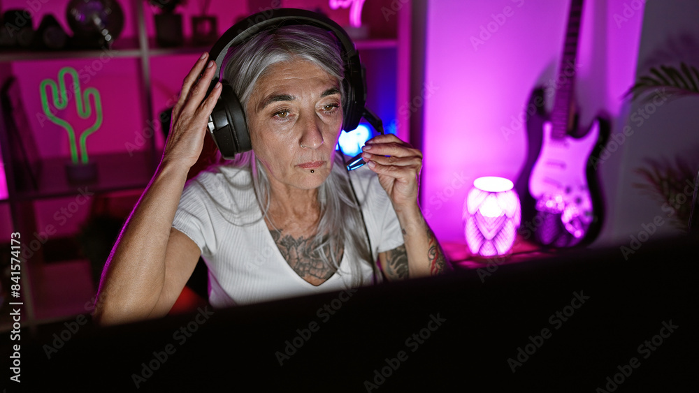Mature, grey-haired woman gamer, in serious gaming mode, streaming live in the heart of the night from her cozy home gaming room, sporting headphones and a mic Mature, grey-haired woman gamer, in serious gaming mode, streaming live in the heart of the night from her cozy home gaming room, sporting headphones and a mic