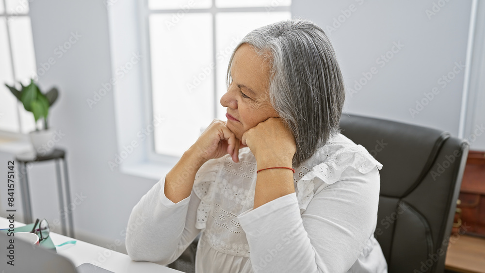 Serene senior woman resting chin on hands at her modern office space, exuding professionalism and calmness. Serene senior woman resting chin on hands at her modern office space, exuding professionalism and calmness.