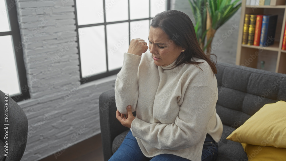 A middle-aged hispanic woman looking pensive indoors, seated on a couch in a cozy living room setting. A middle-aged hispanic woman looking pensive indoors, seated on a couch in a cozy living room setting.