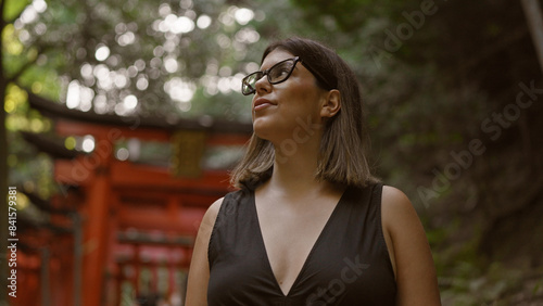 Confidently posing at fushimi inari taisha, a beautiful hispanic woman wearing glasses, smiles broadly, looking around, captivated by the stunning kyoto temple surroundings. photo