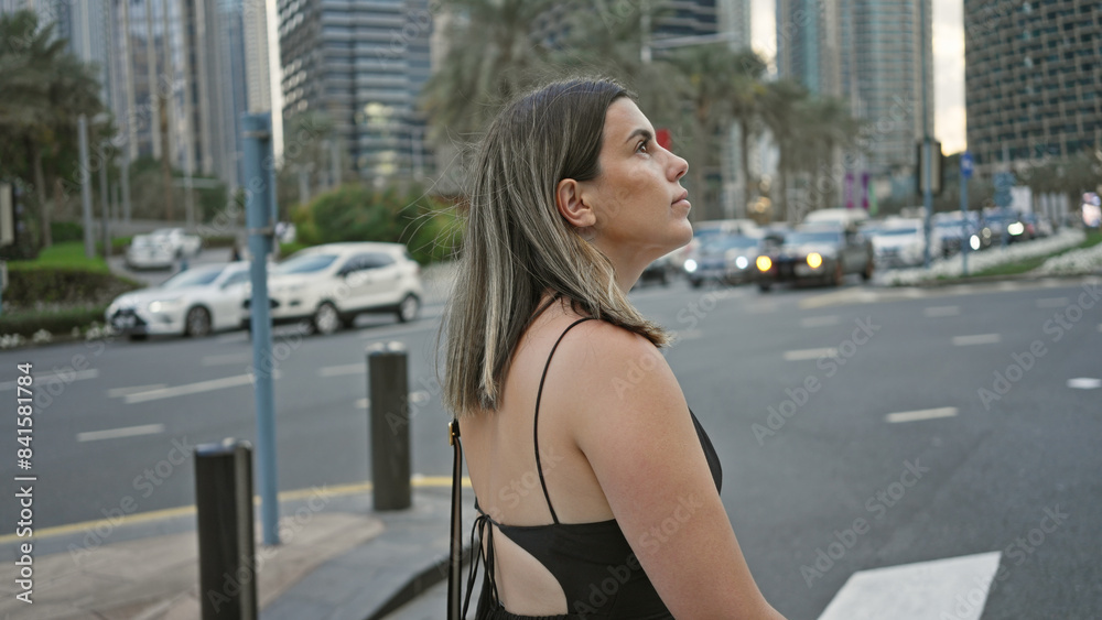 A contemplative young woman with brunette hair standing in modern downtown dubai amidst skyscrapers.