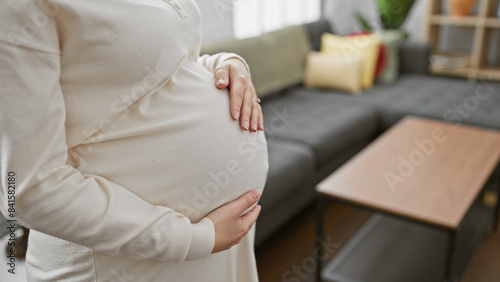 Close-up of a pregnant hispanic woman cradling her belly while standing in a cozy living room setting.