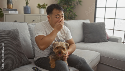 A young chinese man is sitting indoors on a couch in a living room while petting his dog, appearing surprised as he watches something intently.