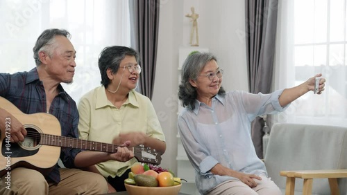 Handheld shot of happy elderly friends in a living room. One plays the guitar while another snaps a selfie, preserving their cherished moments of music and companionship.