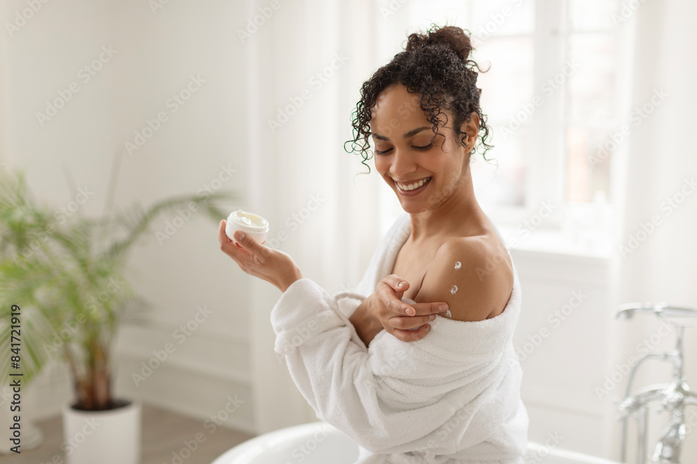 Attractive black lady applying moisturizer on bare shoulders, moisturizing skin, sitting in modern bathroom interior. Skincare cosmetics concept Attractive black lady applying moisturizer on bare shoulders, moisturizing skin, sitting in modern bathroom interior. Skincare cosmetics concept