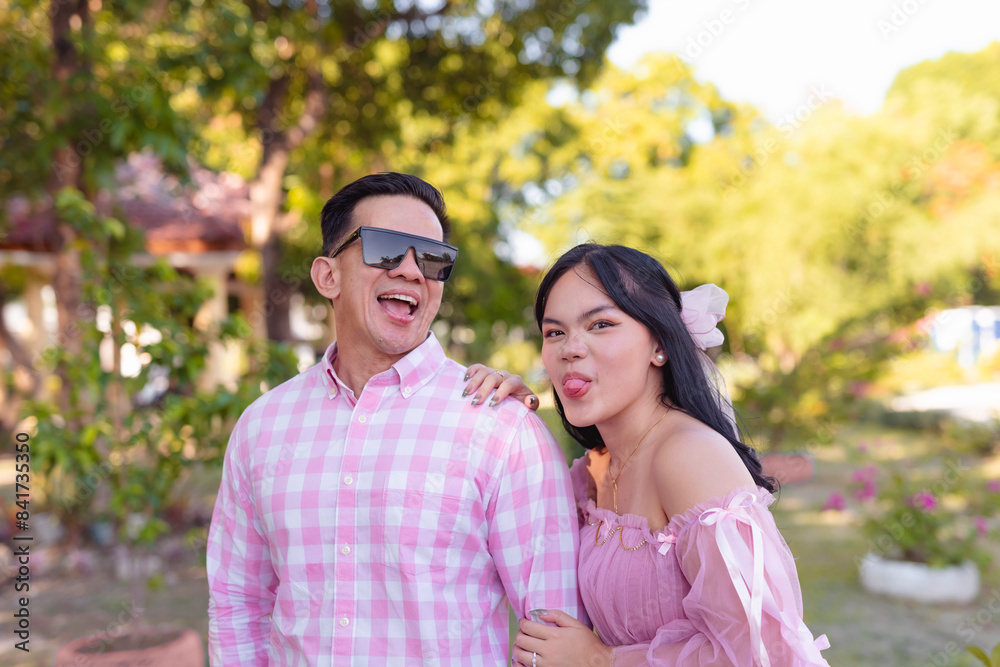 A handsome youthful asian man horsing around with his teenage daughter in pink clothes celebrating Father's Day outdoors at the park. A handsome youthful asian man horsing around with his teenage daughter in pink clothes celebrating Father's Day outdoors at the park.