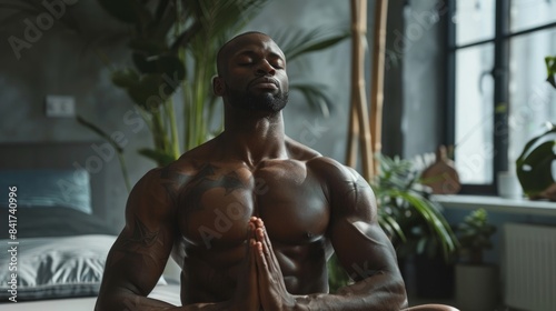 A muscular man sits on a bed in his home, meditating with his eyes closed. He is shirtless, and the image is shot from a low angle.