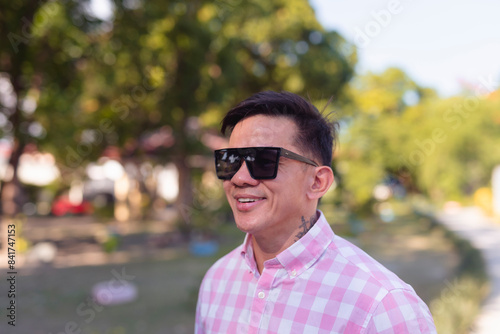Outdoor portrait of a youthful looking Asian man in his mid 40s, with a neck tattoo, wearing sunglasses and smiling in a park on a sunny day.