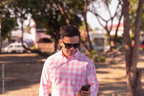 Youthful looking Asian man in his mid 40s with a neck tattoo, wearing sunglasses and a pink plaid shirt, and using his smartphone in an outdoor park setting.
