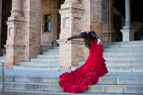 Beautiful woman dancing flamenco in a square in Seville, Spain. She is wearing a typical red and black gypsy dress and dancing flamenco with a lot of art. The dress hangs down the steps of the square.