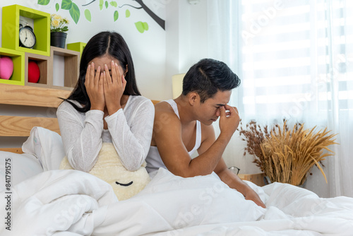 A couple is sitting on a bed looking upset. The woman is crying and the man is holding his head in his hands.