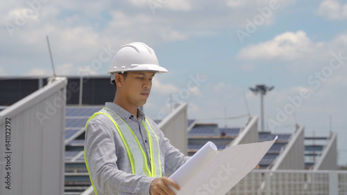An engineer in a hard hat and safety vest reviews solar project plans on a tablet, standing at a site with solar panels.