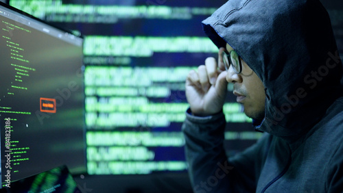 A person wearing a hoodie and glasses closely examines code on multiple screens, suggesting hacking or programming activity in a dark room.