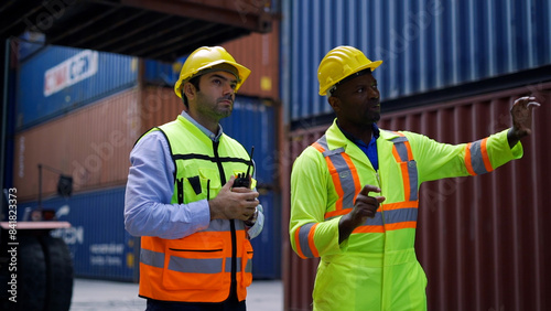 Two workers wearing hard hats and safety vests discuss logistics and operations at a shipping container yard with stacked containers in the background.  