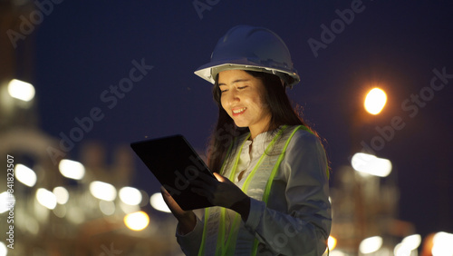 A female engineer wearing a safety helmet and reflective vest uses a tablet for inspection at a brightly lit industrial site at night.