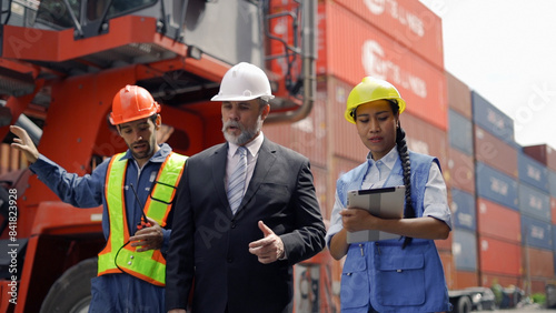 A manager in a suit and hard hat talks with two workers in safety gear at a busy shipping yard with containers in the background.