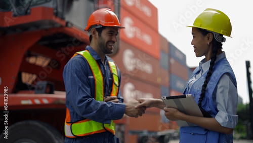 Two workers in safety helmets and vests communicate at a shipping yard, with stacked cargo containers and equipment in the background.