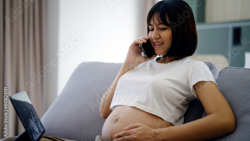 A pregnant woman sits on a couch looking at an ultrasound image of her baby on a tablet screen.