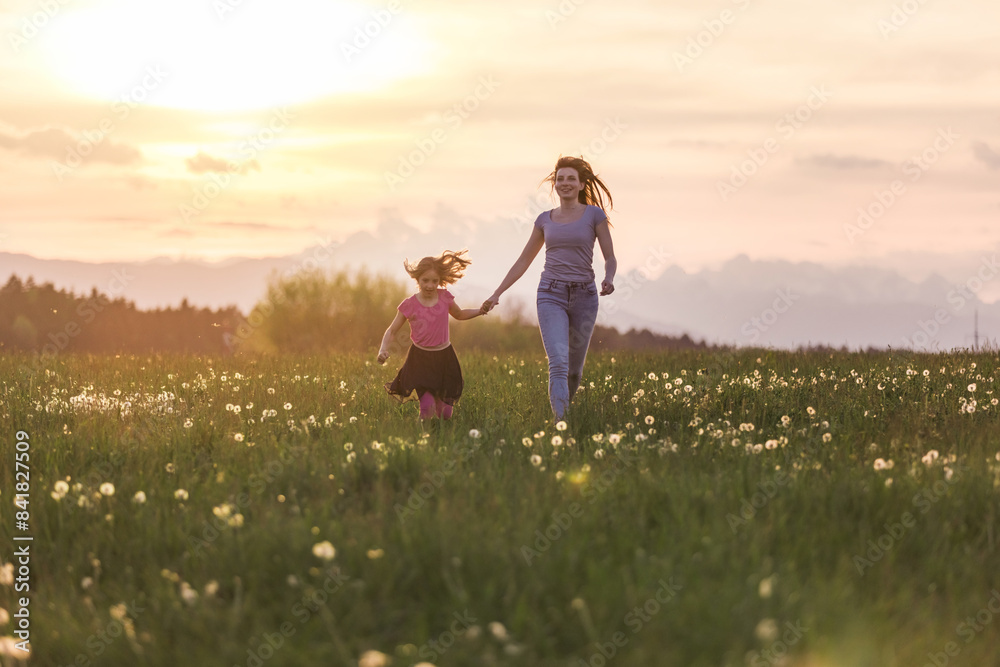 Fototapeta premium Happy and smiling mother and her little daughter running holding hands across a meadow in the beautiful setting sun.