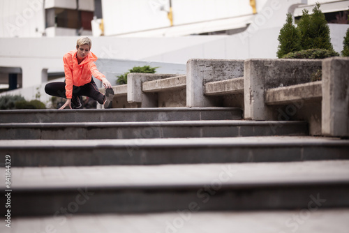 A young woman prepares for a run by stretching on outdoor concrete stairs, wearing a vibrant orange sports jacket and headphones. © Nikola Spasenoski