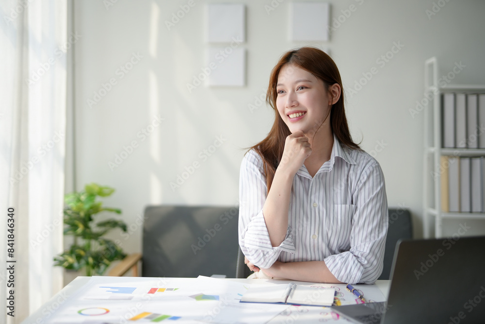 Smiling young woman working at home office desk with laptop, papers, and a plant, illuminated by natural light from window. Smiling young woman working at home office desk with laptop, papers, and a plant, illuminated by natural light from window.