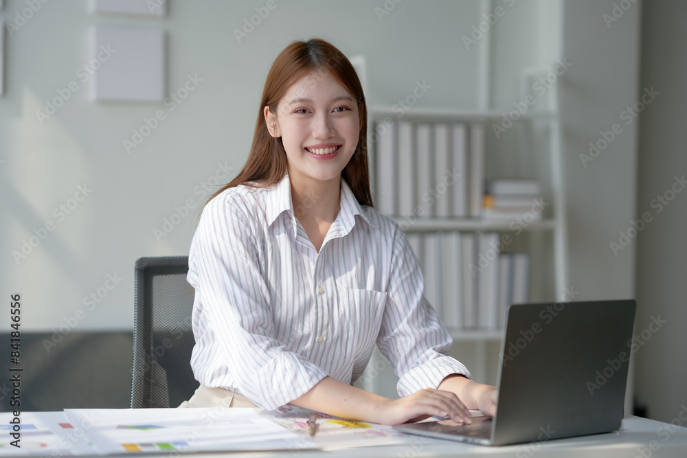Smiling young woman working on a laptop at a modern office desk, looking at the camera with confidence and professionalism.