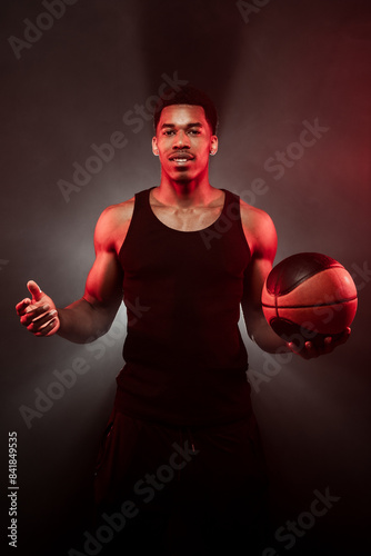 Basketball player side lit with red color holding a ball against hazy smoke fog background. Serious concentrated african american man. © Nikola Spasenoski