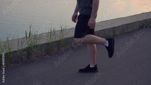 male athlete performs a stretching exercise, pulls his leg with his hand. guy is getting ready for a run on the riverbank at sunset in an urban environment. photo